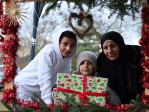 Zwei Jungen und Mutter. Kleiner Junge mit Mütze in der Mitte hat großes Geschenk in den Händen. Jugendlicher trägt weißen Hoodie, Mutter mit dunkler Kleidung und Kopftuch. Alle schauen in die Kamera.