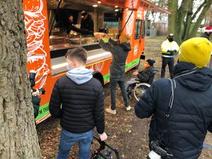 Orangener Food Truck. Menschen in Winterjacken, Mützen und Masken stehen davor. Einer nimmt gerade Essen aus dem Truck entgegen. Ein Rollstuhlfahrer steht neben der Person und streckt die Hände aus um das Essen entgegenzunehmen.