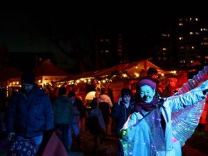 Beleuchtete Buden, Gäste auf dem Markt. Rechts eine Frau mit roter Nase und glitzernden leuchtenden Flügeln bewegt sich durch die Dunkelheit.