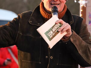 Draußen. Moderator mit Mikro in der Hand. Er trägt eine lila Mütze und dunkle Kleidung.