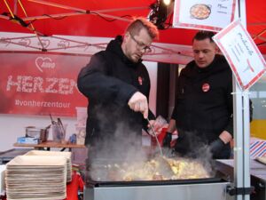 Zwei Männer in einem AWO Pavillon wo es Bratwurst und ein vegetarisches Pfannengericht gibt.. Bratwürste liegen auf dem Grill. Einer der Männer bereitet das Pfannengericht zu.