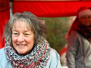 Frau lächelt in die Kamera. Sie trägt einen dicken Schaal daran ist ein Button der Von Herzen Tour befestigt. Im Hintergrund eine Frau mit Weihnachtsmütze unscharf.
