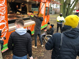 Orangener Food Truck. Menschen in Winterjacken, Mützen und Masken stehen davor. Einer nimmt gerade Essen aus dem Truck entgegen. Ein Rollstuhlfahrer steht neben der Person und streckt die Hände aus um das Essen entgegenzunehmen.