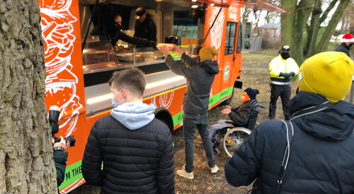 Orangener Food Truck. Menschen in Winterjacken, Mützen und Masken stehen davor. Einer nimmt gerade Essen aus dem Truck entgegen. Ein Rollstuhlfahrer steht neben der Person und streckt die Hände aus um das Essen entgegenzunehmen.