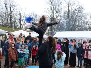 Ein Mädchen ballanziert auf einem Bein auf den Händen eine Frau. Das linke Bein streckt das Mädchen nach hinten, die Arme sind zur Seite ausgestreckt. Sie hat einen Pompon in jeder Hand, blau und Silber. Zuschauer stehen im Halbkreis darum.