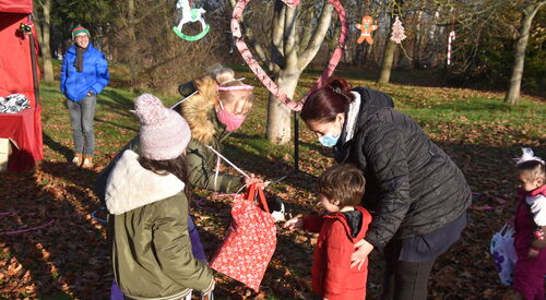 Draußen, Laub auf Wiese, Bäume. Zwei Kinder, Mädchen und Junge in Winterkleidung stehen mit zwei Frauen die Maske tragen zusammen. Eine Frau hat roten Beutel mit etwas darin in der Hand und hält es dem kleinen Jungen hin.
