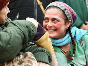 Frau mit zum Clown geschminktem Gesicht hockt neben alter Frau im Rollstuhl. Lächelt breit und reicht der alten Frau ihre Hand.