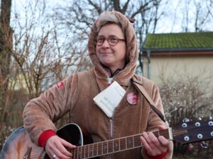 Eine Frau, die Gitarre spielt. Die grauen Haare unter einer brauen Kapuze, runde Brille mit braunen Gestell. Sie trägt eine braune Jacke mit Button. Die Gitarre ist braun-schwarz.