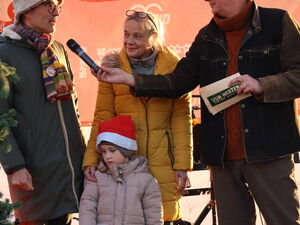Moderator hält einem Mann ein Mikro hin. Zwischen ihnen steht eine blonde Frau die ein Kind mit roter Weihnachtsmütze vor sicch stehen hat.
