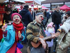 Draußen. Buntes Weihnachtsmarkttreiben. Im Vordergrund eine Clownin mit Seifenblasen und ein Clown spielt auf einer Ukulele, beide haben rote Nasen und sind geschminkt.