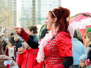 Fee Lilli mit rotem Kleid, Krone und glitzernden Flügeln hält einen Zauberstab vor sich. Sie ist von der Seite fotografiert. Drum rum viel große und kleine Gäste.