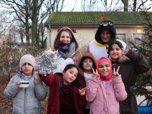 5 Kinder, dahinter 2 Erwachsene. Frau im Elchkostüm, Mann im Pinguin Kostüm. 3 Mädchen, 2 Jungen, alle tragen Winterkleidung. Mädchen in Rosa Winterjacke und pinkem Kopftuch zeigt mit beiden Händen das Victory-Zeichen. Alle lächeln in die Kamera.