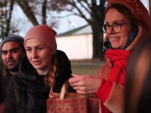 Draußen. 2 Frauen, dahinter ein Mann. Sie tragen Winterkleidung. Frau im Vordergrund, mit rotem Schal, Brille mit schmalen schwarzen Rahmen, hält eine Weihnachtstüte vor sich. Die Frau daneben trägt eine blassrosa Mütze und schwarze Winterjacke.