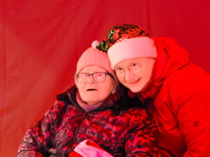 In der Fotobox. Roter Hintergrund. Zwei Menschen. Ältere Frau im Rollstihl und Frau mit Weihnachtsmütze und roter Jacke posieren für ein Foto.