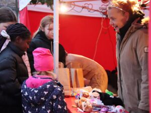 Vier Mädchen, eines davon schwarz stehen an einem Stand wo es kleine Geschenke gibt, z.B. kleine Kuscheltiere.Eine Frau im Stand lächel die Kinder an.